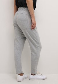 Persoon in lichtgrijze joggingbroek en witte sneakers, staat tegen een effen witte muur, met een hand op de heup.