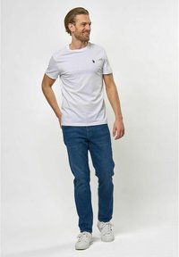 Bela bomba majica iz bombaža s kratkimi rokavi in majhnim mornarško modrim logotipom na prsih, v kombinaciji s temno modrimi džinsom in belimi supergami.