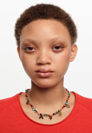 Persona joven con cabello corto y rizado que lleva un collar colorido de cuentas con varios amuletos dorados y una camisa roja.