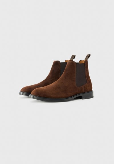 Botine Chelsea din suede maro, cu panouri elastice pe laterale și cârlige pentru a fi ușor de încălțat. Vârf rotunjit și talpă din cauciuc negru. Toc plat pentru stabilitate.