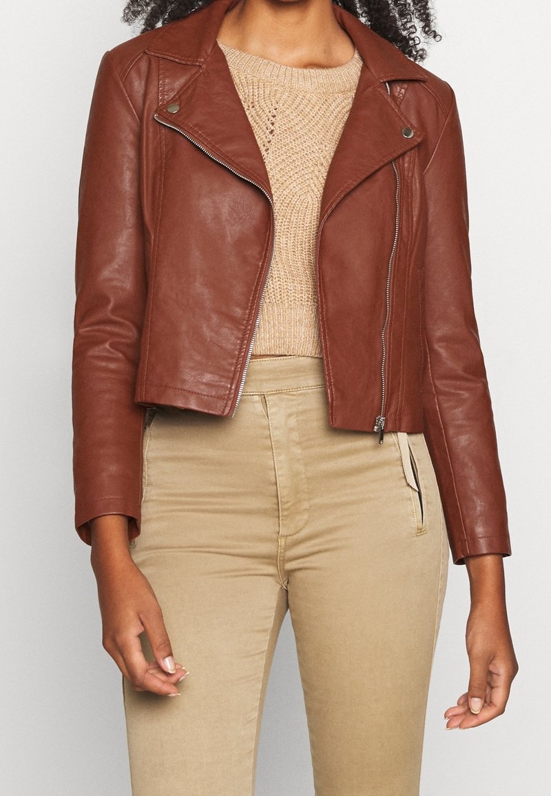Kobieta w brązowej skórzanej kurtce o kroju crop, założonej na beżowy sweter dziany i beżowe spodnie z wysokim stanem, stoi na tle jednolitym.