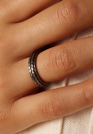 Zwart gevlochten metalen ring rustend op een hand met een gestructureerde huid; zachte stofachtergrond met een subtiele lichte kleur.