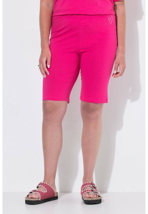 Personne portant un short rose vif jusqu'aux genoux avec un petit détail cœur blanc sur le côté gauche et des sandales roses assorties avec une couture blanche.