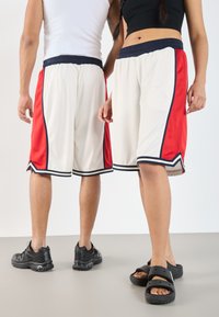 Shorts de sport blancs avec des panneaux latéraux rouges, une taille et des finitions bleu marine. Présentent une coupe ample et une texture en mesh. Marquage notable sur les chaussures.