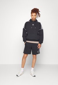 Felpa nera con strisce bianche, tasca kangaroo e logo. Abbinata a pantaloni corti neri coordinati e sneakers bianche, con un top a strati.