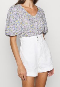 Haut floral à manches bouffantes en lavande, jaune et bleu avec un col en V ; associé à un short blanc taille haute avec fermeture à boutons et poches.