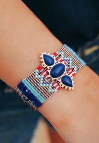 Bracelet en perles avec une pierre précieuse bleue au centre, motifs tissés complexes en rouge, bleu et turquoise, avec des accents métalliques.