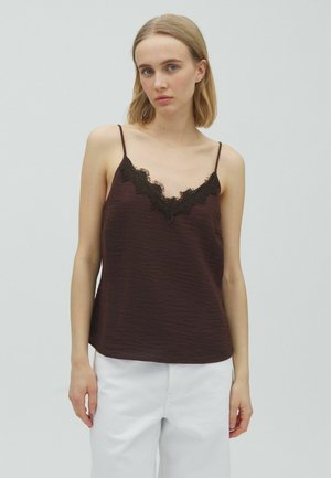 Tânără cu păr blond, purtând o camisă maro închis cu dantelă și pantaloni albi, stând pe un fundal deschis și simplu.