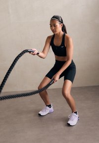 Soutien-gorge de sport noir et shorts, chaussures de sport bicolores violettes, tenant une corde de bataille épaisse et texturée, dans un cadre intérieur de tons neutres.