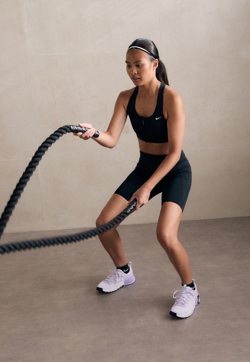 Reggiseno sportivo nero e pantaloni corti, scarpe da ginnastica atletiche bicolori viola, in mano una corda da battaglia spessa e texturizzata, in un ambiente interno dai toni neutri.
