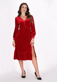 Rochie din catifea roșie cu mâneci lungi, de tip V, cu nasturi pe față și tiv volanat. Prezintă o deschidere laterală și un design strâmt în talie.
