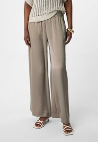 Beige wijde broek van lichtgewicht stof met een elastische tailleband. Gecombineerd met witte sandaaltjes met bandjes, wat een ontspannen pasvorm laat zien.