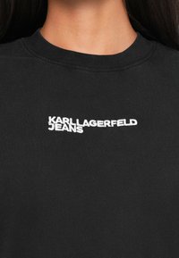Černé bavlněné tričko s kulatým výstřihem a bílým vyšívaným nápisem "KARL LAGERFELD JEANS" na přední straně. Hladká textura.