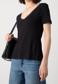 Eine Frau trägt ein schwarzes T-Shirt mit Spitzenbesatz im V-Ausschnitt, blaue Jeans, hält eine schwarze Umhängetasche und trägt goldene Ringe an der rechten Hand.