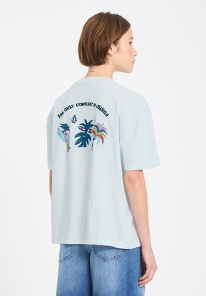 Personne aux cheveux courts bruns portant un T-shirt bleu clair avec des motifs colorés abstraits de plantes et le texte "LA SEULE CONSTANTE EST LE CHANGEMENT."