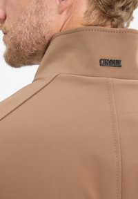 Beige jas met hoge kraag, voorzien van een zwart "CINQUE" logo. Soepele textuur en zichtbare stikseldetails op de naden.
