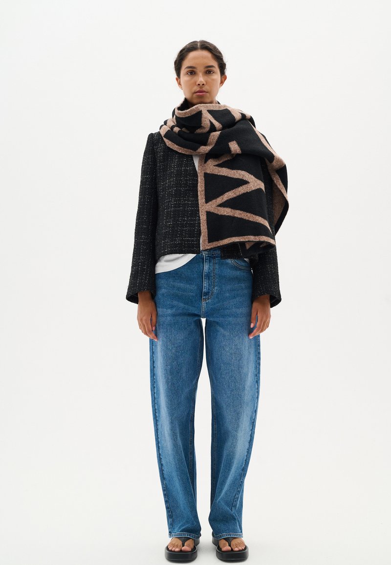 Giacca corta in tweed nero sopra una camicia bianca, abbinata a jeans blu a vita alta. Un grande sciarpa a motivi neri e marroni è drappeggiata in modo casual.