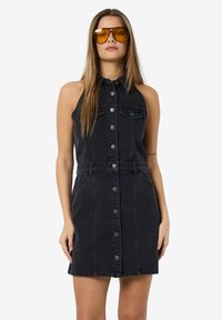 Zwart denim mouwloos jurk met een knoopsluiting aan de voorkant, twee zijzakken, een aangesloten taille en een rechte zoom. Model met gele zonnebril.