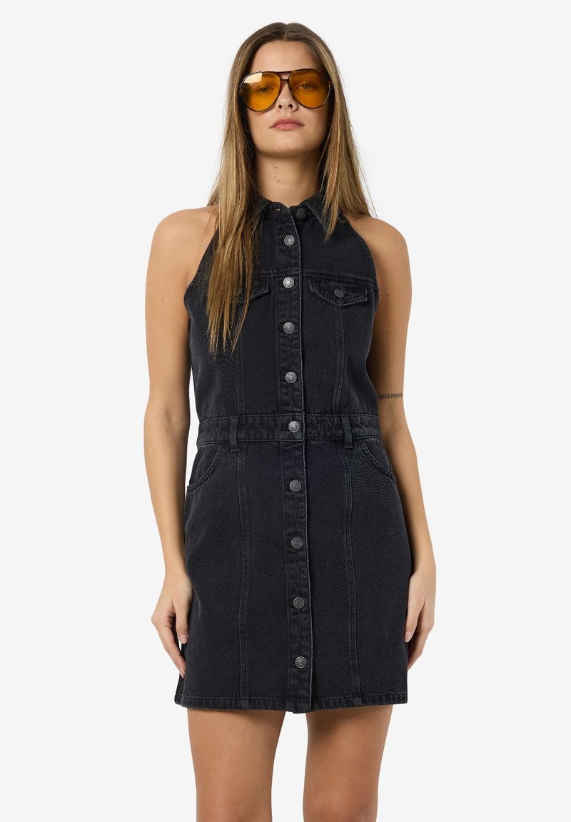 Zwart denim mouwloos jurk met een knoopsluiting aan de voorkant, twee zijzakken, een aangesloten taille en een rechte zoom. Model met gele zonnebril.