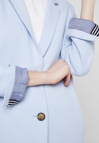 Blazer azzurro chiaro con una texture liscia, rever a punta e polsini a strisce. Un singolo bottone tondo è centrato sul davanti.