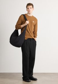 Καφέ ριχτό μακρυμάνικο μπλουζάκι με μικρό λογότυπο, μαύρη κεντητή τσάντα hobo, μαύρο φαρδύ παντελόνι και μαύρα παπούτσια σε ουδέτερο φόντο.