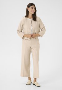 Blazer bej scurt cu mâneci 3/4 și butoane decorative; pantaloni largi asortati dintr-un material ușor; stilizat cu loafers bej.