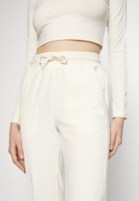 Bílý krátký top s dlouhým rukávem ve spojení s lehkými krémovými tepláky. Tepláky mají stahovací pás a decentní logo na boku.