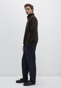 Brauner Fleece-Pullover mit hohem Kragen, kombiniert mit locker sitzenden dunkelblauen Jeans und schwarzen Schnürschuhen. Schlichter Hintergrund.