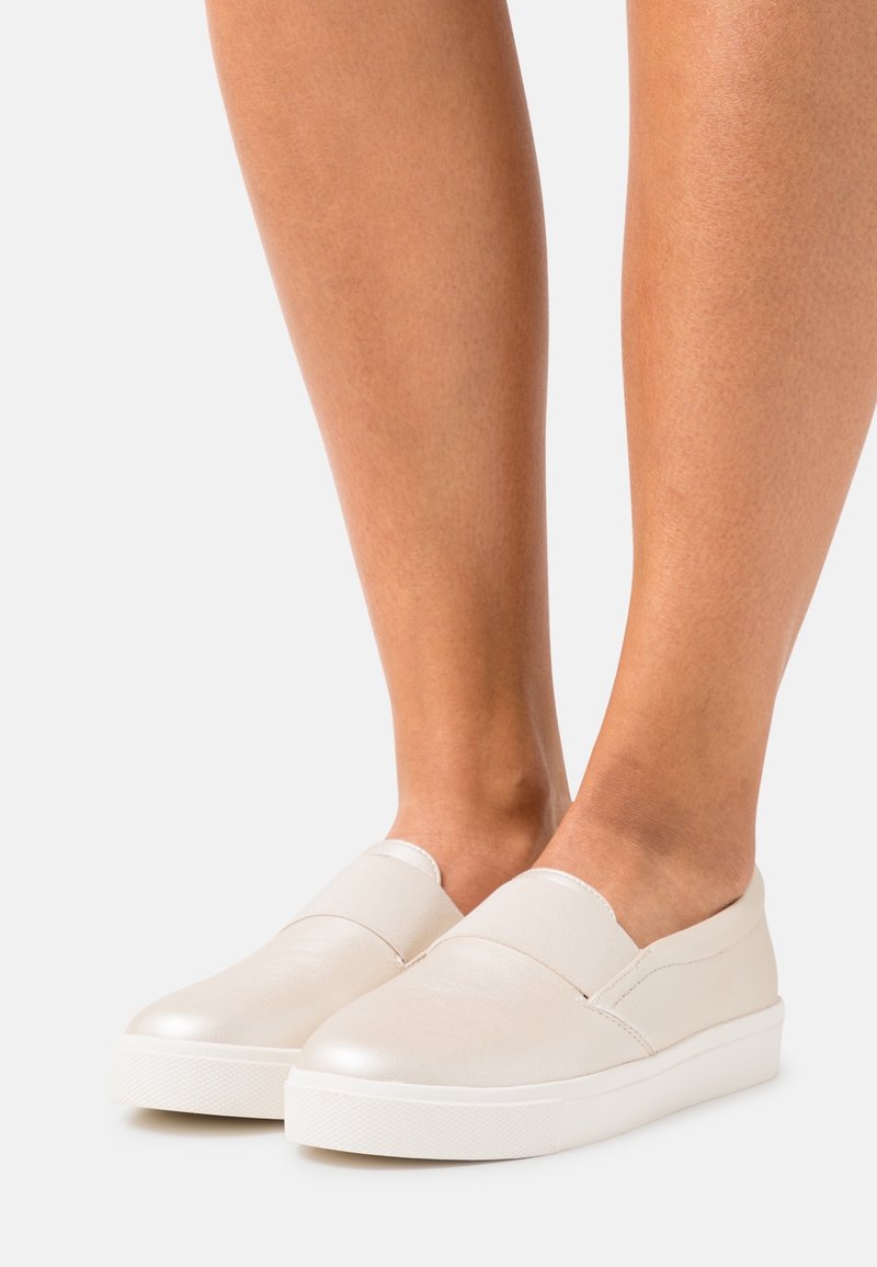 Jambes portant des baskets slip-on écrues avec semelles blanches épaisses sur un fond clair uni.