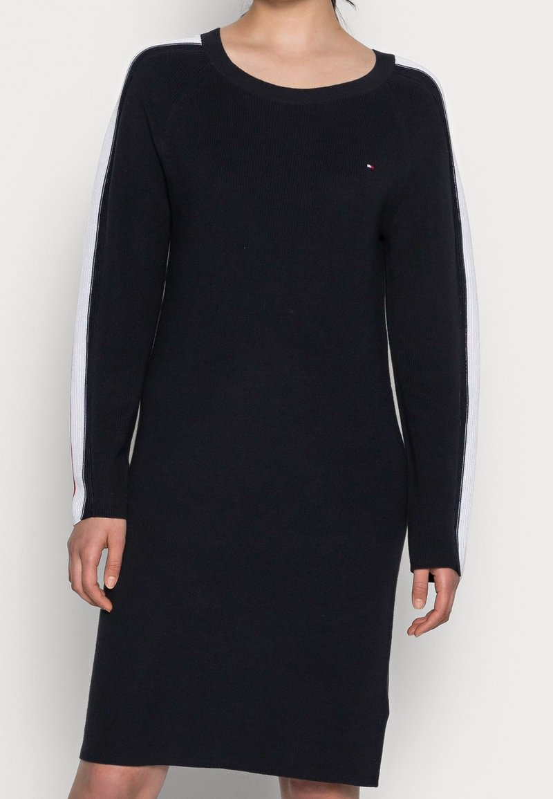 Robe en maille noire à manches longues avec des rayures blanches le long des manches et un petit logo rouge, blanc et bleu sur le haut de la poitrine.