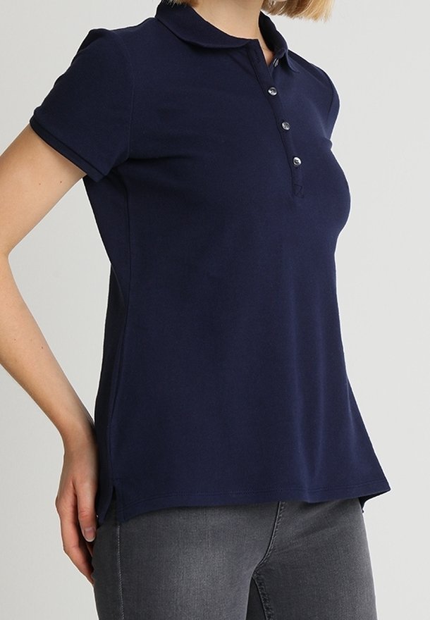 Femme portant un polo bleu marine à manches courtes avec des boutons et un pantalon gris ajusté, debout devant un fond clair uni.