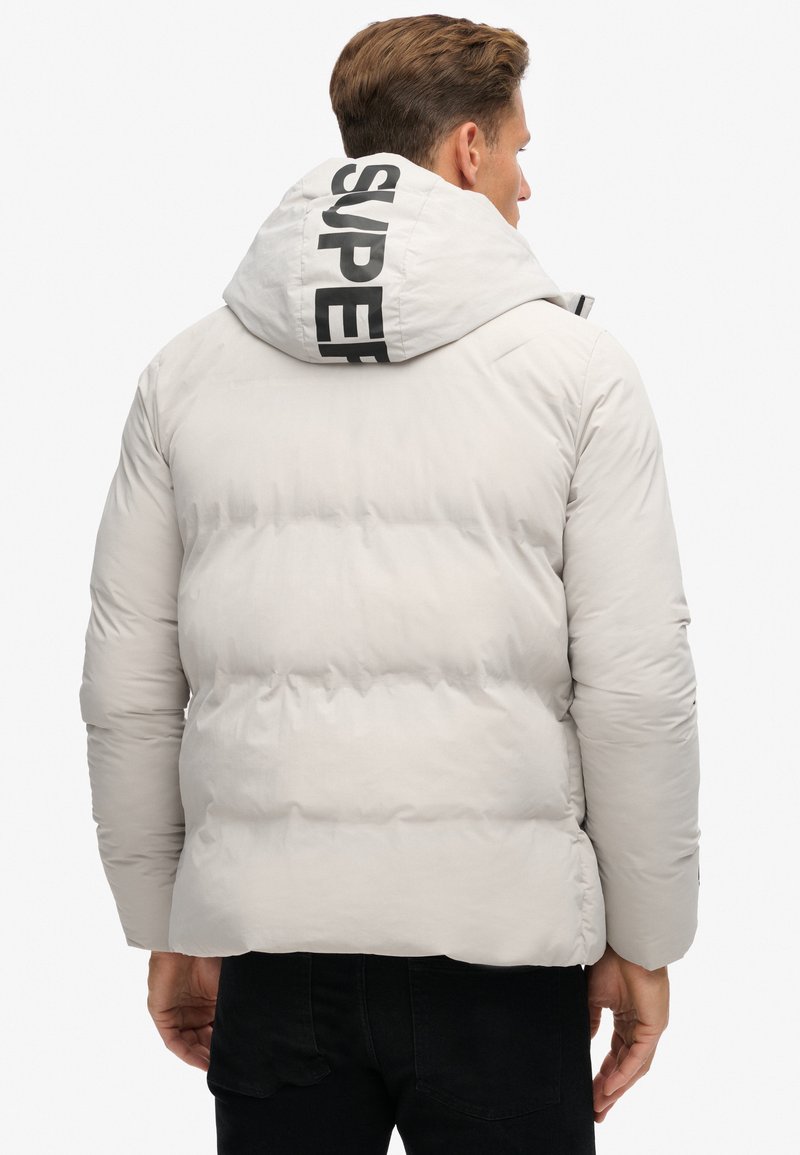 Dikke, lichtgrijze jas met zwart "SUPER" logo op de capuchon, ritsluiting, horizontale vulling en elastische manchetten. Gelaagde afwerking en eigentijds ontwerp.