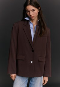 Mlada ženska z dolgimi temnimi lasmi nosi preveliko temno rjavo srajco prek svetlo modre srajce in svetlo opranih kavbojk, stoji na temnem ozadju.