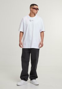 White cotton oversized t-shirt with brand logo, paired with dark gray wide-leg pants and white sneakers. Character wears dark sunglasses.