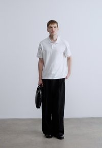 Fehér, galléros rövid ujjú póló, laza fekete nadrággal párosítva. A modell fekete táskát visel, és semleges háttér előtt áll.