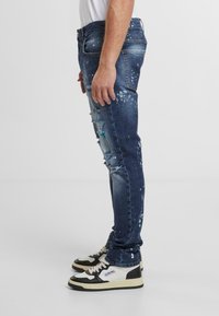 Mörkblå denimjeans med slitna detaljer, ljus färgsplatter och en slim fit-design. Kombinerat med svarta och vita sneakers.