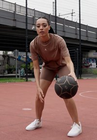 T-shirt de sport marron et shorts, baskets blanches, tenant un ballon de basket noir à motifs étoiles sur un terrain rouge avec un fond clôturé.