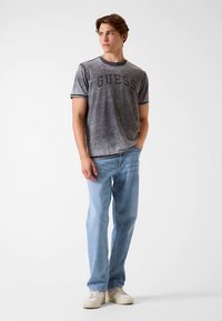 T-shirt gris à col rond avec l'inscription "GUESS" en lettres foncées, assorti à un jean en denim bleu clair et des baskets blanches.