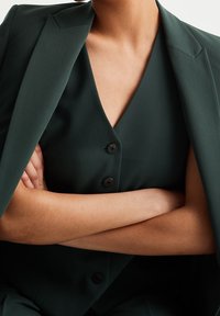 Blazer vert ajusté avec des revers nettes, superposé à un haut sans manches assorti présentant un col en V et trois boutons foncés. Texture de tissu lisse.