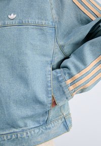 Denim jakna u svijetloplavoj boji s bež trostrukim uzorkom na rukavima. Ima malu oznaku s logotipom i klasični ovratnik.