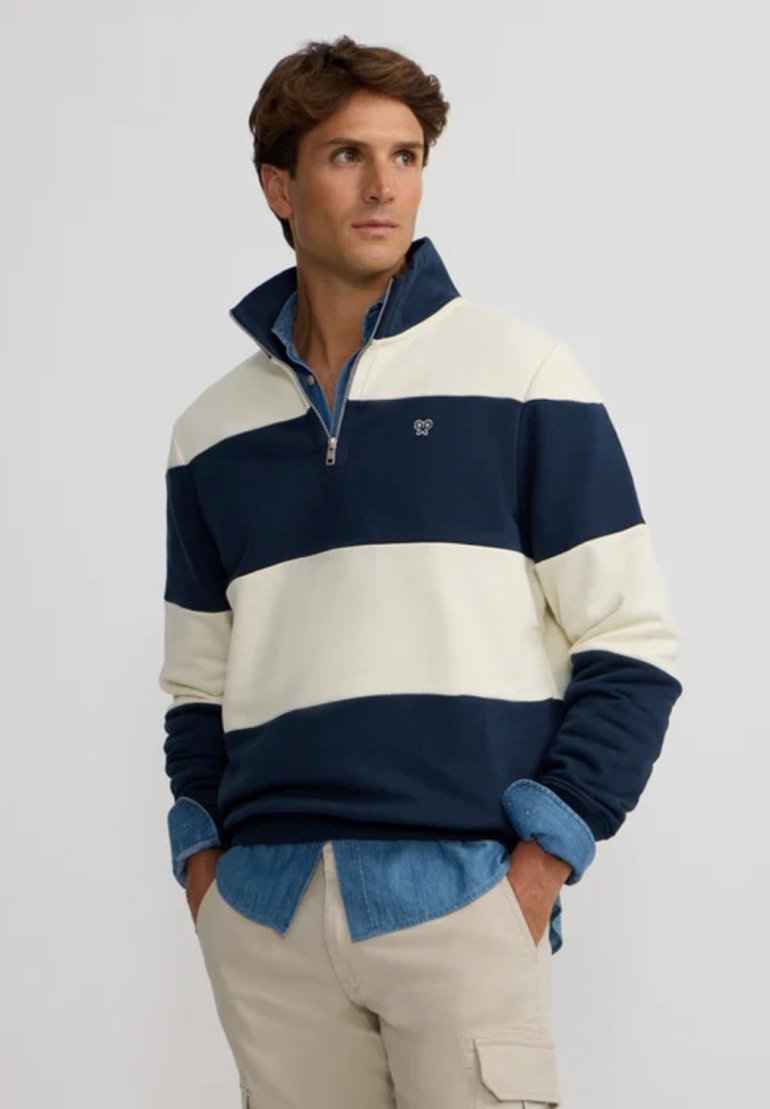 Pull à rayures bleu et crème avec un col haut et une fermeture éclair, arborant un logo sur la poitrine, fabriqué dans un tissu doux.