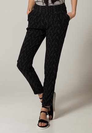 Black patterned trousers with an elastic waistband, side pockets, and tapered legs, paired with black sandals and a gray graphic top.