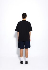 Czarny oversize t-shirt z krótkimi rękawami, w połączeniu z ciemnymi dżinsowymi szortami. Całość uzupełniają białe skarpety i czarne buty. Proste tło.