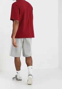 Man wearing a red T-shirt, gray athletic shorts with white stripes, white socks, and gray Adidas sneakers walking away on a white floor.