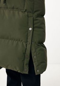 Dunkelgrüne gesteppte Jacke, Seitenansicht mit geöffneten und geschlossenen schwarzen Druckknöpfen, über schwarzer Hose, auf weißem Hintergrund.