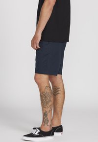 Uomo che indossa pantaloni corti blu navy, t-shirt nera e sneakers nere con suola bianca, mostrando tatuaggi su entrambe le calve contro uno sfondo grigio uniforme.