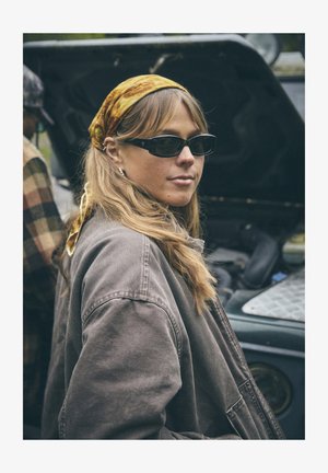 Mulher usando óculos de sol pretos e um casaco castanho largo, com cabelo longo e ondulado e um lenço amarelo texturizado amarrado na cabeça.