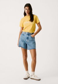 Gelbes Baumwoll-T-Shirt mit weißem "Red Bull Racing"-Logo, kombiniert mit hoch taillierten blauen Jeansshorts mit ausgefranstem Saum und Taschen. Weiße Sneakers.