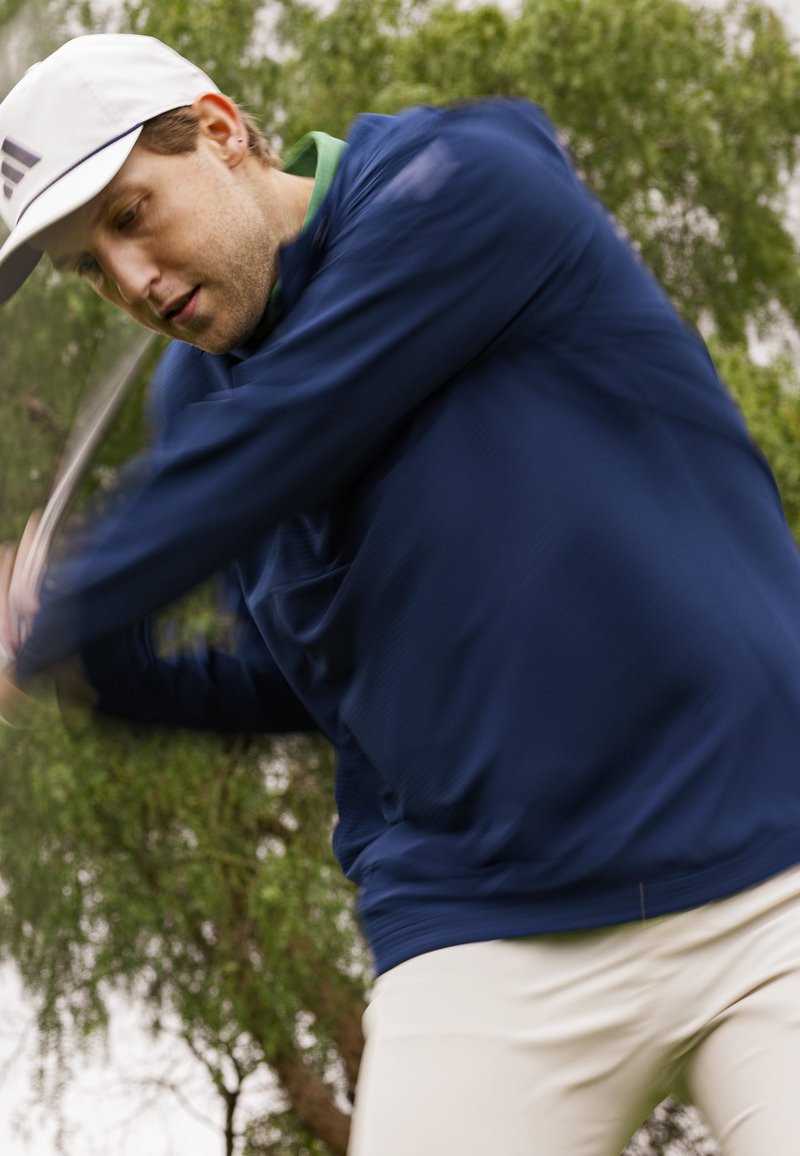 Golfista w granatowej, długiej kurtce i jasnych spodniach, wykonujący zamach kijem pośród zielonej roślinności w rozmytym ujęciu akcji.