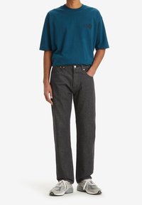 Man draagt een turkooizen Levi’s T-shirt, donkergrijze rechtgesneden spijkerbroek en grijze en witte sneakers, staat met één hand in de zak.